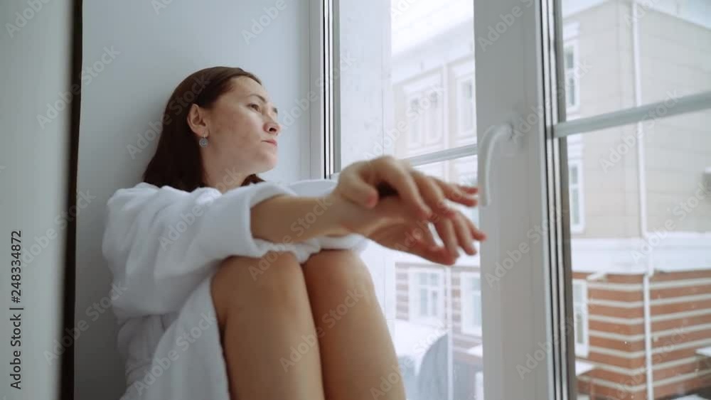 A depressed woman in a white coat sits on the windowsill and looks out into the street. It is snowing outside. A sad woman is sitting on the windowsill. A depressed woman in a white coat sits on the windowsill and looks out into the street. It is snowing outside. A sad woman is sitting on the windowsill.