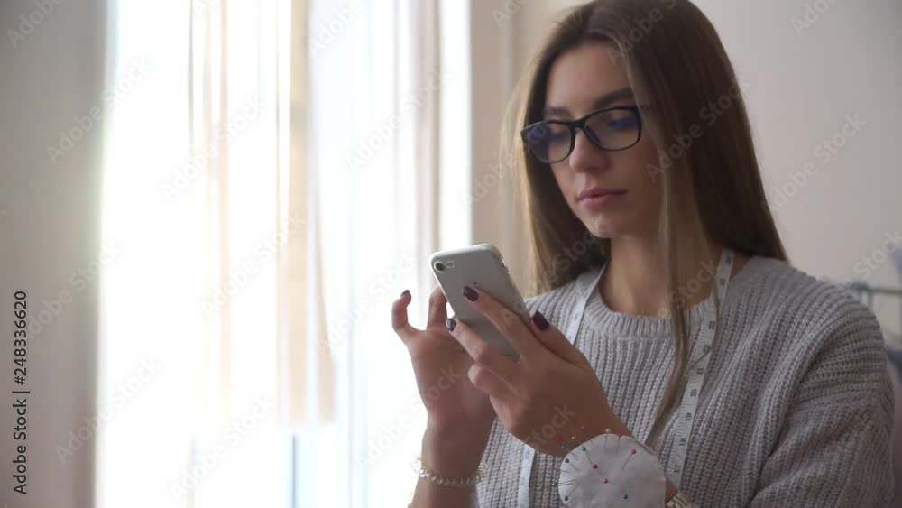 Young Russian girl seamstress with glasses. Looking in the phone against the window. From where the sun shines. Young Russian girl seamstress with glasses. Looking in the phone against the window. From where the sun shines.