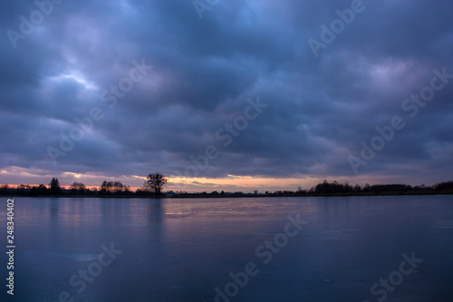 frozen lake, trees on the shore and clouds in the sky after sunset © darekb22
