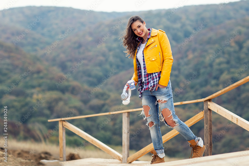 Naklejka premium Adorable long hair girl standing at the wooden bridge with forest on the background with headphones and coffer to go on hand