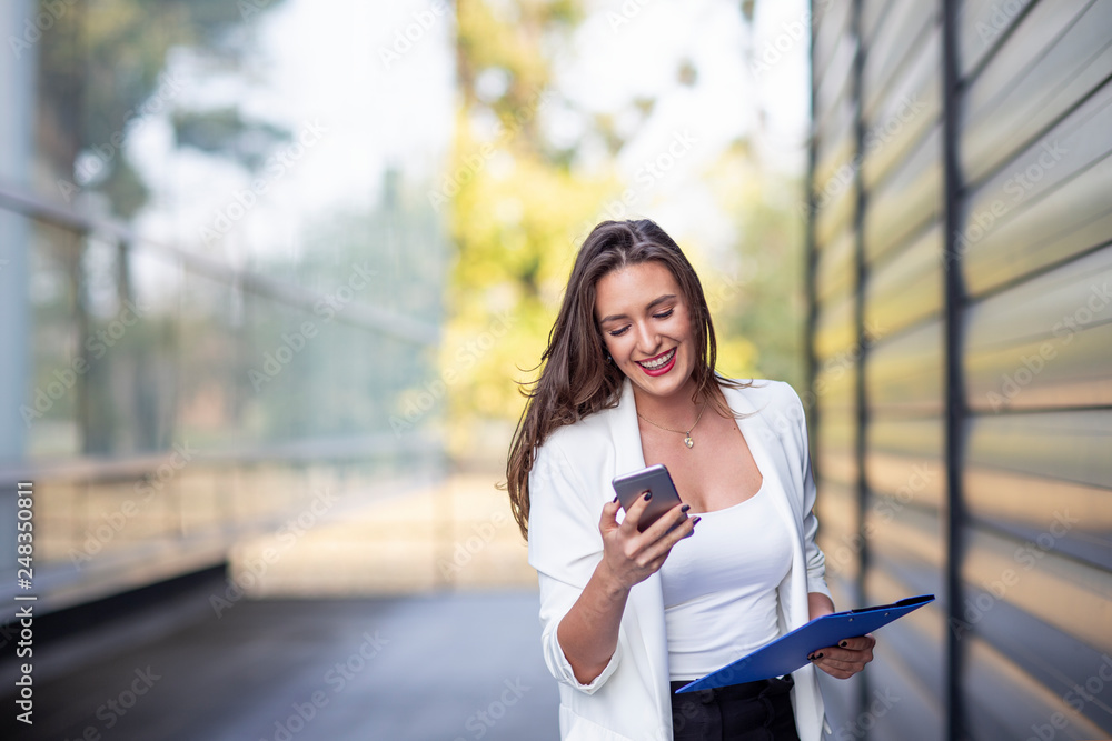 Fototapeta premium Young long hair woman in white dress using mobile phone with documents on hand near the office