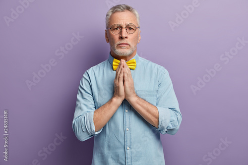People, age and spirituality concept. Serious mature wrinkled man keeps hands pressed together, believes in good luck, wears denim elegant shirt and yellow bowtie, isolated over purple background