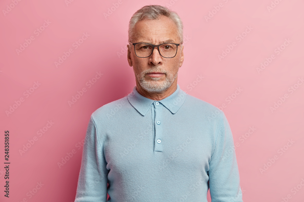 Headshot of grey haired unshaven man wears optical glasses, has serious expression, dressed in casual blue jumper with collar, thinks over something models over rosy background. Thoughtful grandfather Headshot of grey haired unshaven man wears optical glasses, has serious expression, dressed in casual blue jumper with collar, thinks over something models over rosy background. Thoughtful grandfather