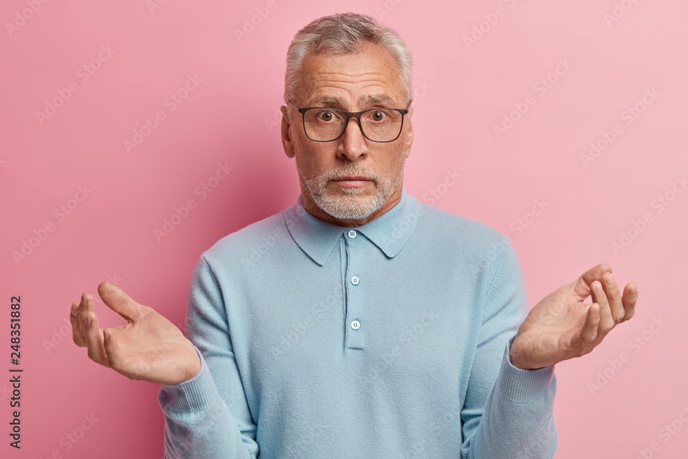 Indoor shot of hesitant elederly man with grey short hair, thick beard, spreads hands in bewilderment, wears spectacles and light blue sweater, makes hard choice or decision. Uncertainty concept Indoor shot of hesitant elederly man with grey short hair, thick beard, spreads hands in bewilderment, wears spectacles and light blue sweater, makes hard choice or decision. Uncertainty concept