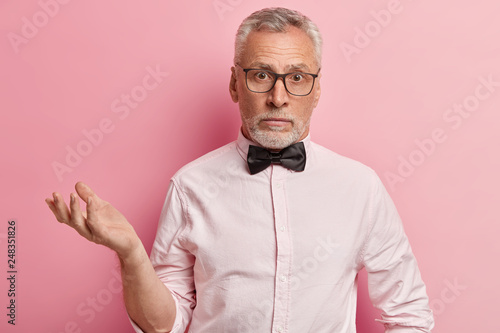 Indoor shot of puzzled mature man with thick grey stubble, wears spectacles, dressed in elegant shirt with black bowtie, gestures in hesitation, poses over rosy background. Doubtful grandfather