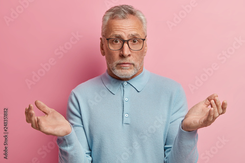 Fotografia Indoor shot of hesitant elederly man with grey short hair, thick beard, spreads hands in bewilderment, wears spectacles and light blue sweater, makes hard choice or decision