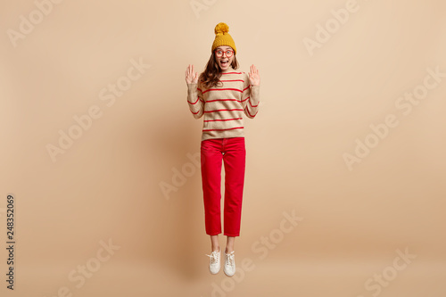 Studio shot of pleased young woman has fun, jumps high, stretches palms forward, wears yellow hat, dressed in sweater and red trousers, isolated over beige background. Wow, its amazing day today