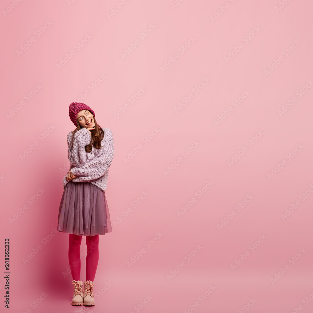 Vertical shot of pleasant looking dreamy smiling woman crosses hands partly over chest, holds chin, looks with enjoyment, dressed in winter outfit, isolated over pink studio wall with copy space aside