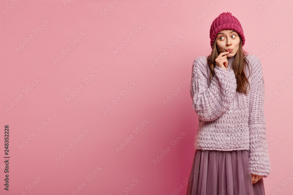 Studio shot of pensive cute young lady focused aside, has folded lips, being deep in thoughts, wears winter clothes, models over rosy studio wall with empty space for your promotional content Studio shot of pensive cute young lady focused aside, has folded lips, being deep in thoughts, wears winter clothes, models over rosy studio wall with empty space for your promotional content