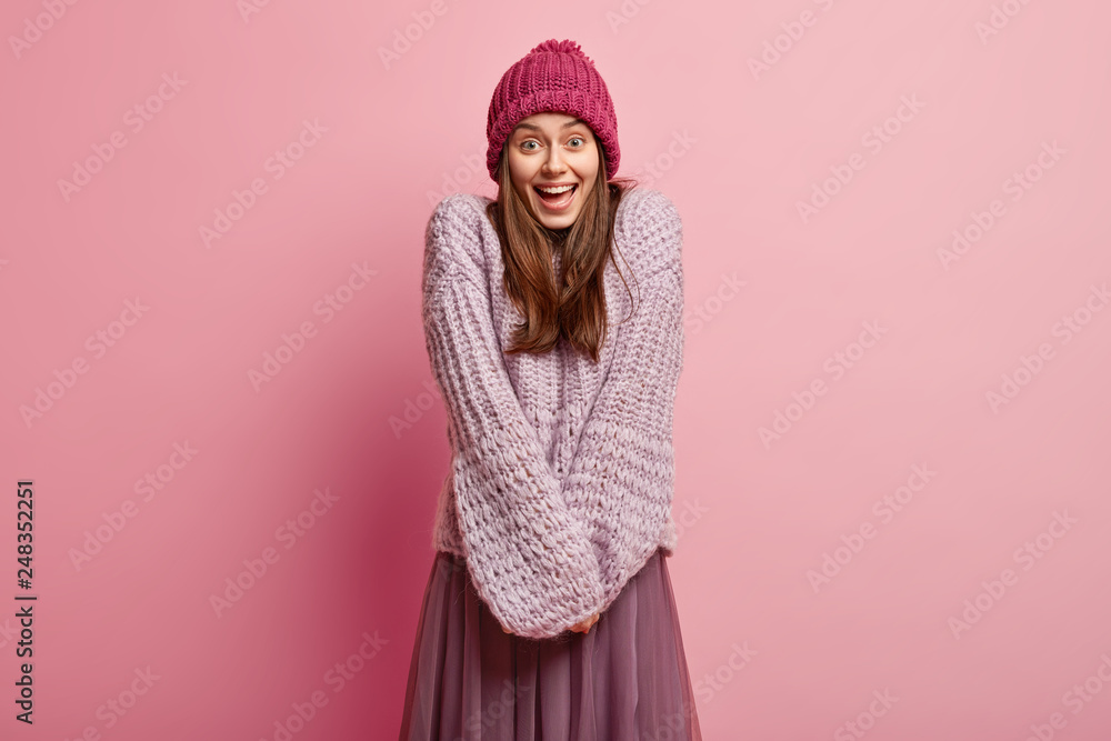 Appealing satisfied woman with shy timid expression, happy look, keeps hands together, wears hat and jumper, recieves good news, isolated over rosy studio wall. People and positive emotions concept. Appealing satisfied woman with shy timid expression, happy look, keeps hands together, wears hat and jumper, recieves good news, isolated over rosy studio wall. People and positive emotions concept.