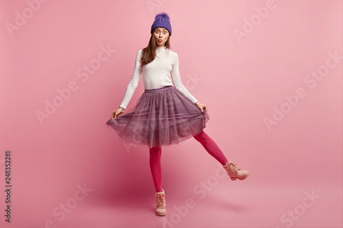 Studio shot of amazed pretty young woman demonstrates her new stylish skirt, wears bright pink tights, raises leg, wears boots, models over rosy background, has eyes popped out, appealing look photo