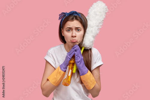 Desperate dissatisfied woman purses lower lip  has upset facial expression  wears headband and casual t shirt  asks for help  uses cleaning detergent and dust brush  isolated over pink background