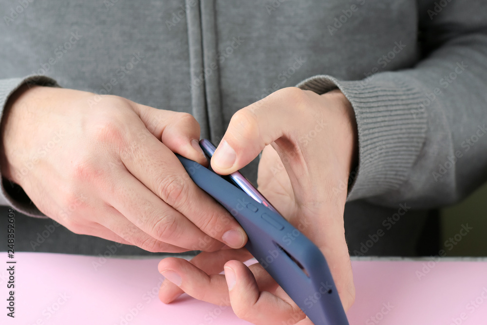 Caucasian man in a gray sweater puts on a new case on mobile phone with selective focus. White male hands are holding a smartphone and putting on a blue cover. Silicone case for digital gadget