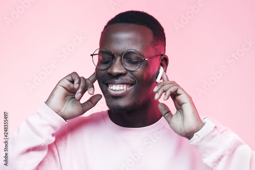 Studio shot of young african man listening to music with wireless earphones isolated on pink background