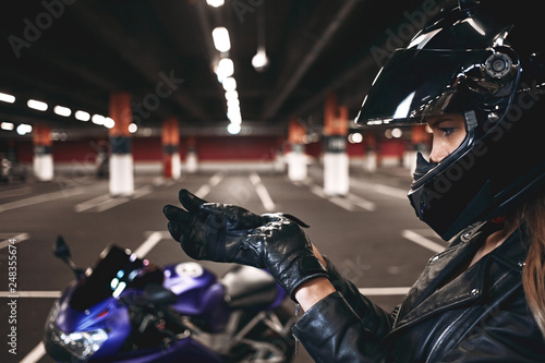 Sideways picture of blonde European girl in fashionable black leather jacket, gloves and protective helmet getting ready before night ride on her motorcycle, posing in deserted parking lot