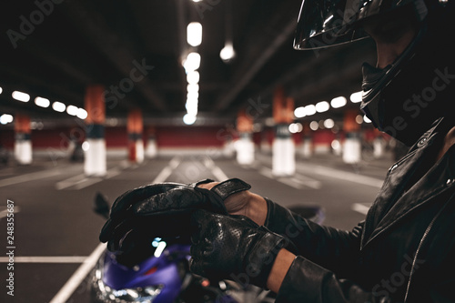Profile portrait of unrecognizable woman biker taking on stylish leather gloves ready for night ride, posing isolated in underground car parking with motorcycle. People, nightlife and extreme sports