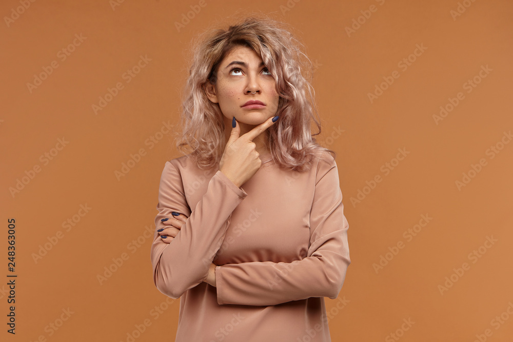 Pensive European female in her twenties making plans for weekend, rubbing chin and looking up, having thoughtful indecisive facial expression. Human emotions, life perception and body language Pensive European female in her twenties making plans for weekend, rubbing chin and looking up, having thoughtful indecisive facial expression. Human emotions, life perception and body language