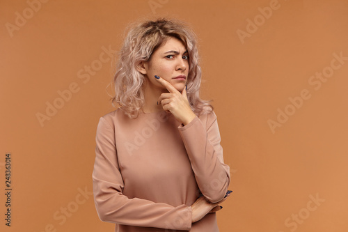 Body language. Portrait of attractive young female with nose ring and voluminous hair touching face and frowning, her look full of distrust and suspicion. Suspicious Caucasian woman posing in studio