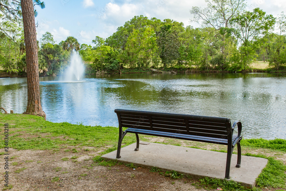 Empty bench next to a lake with a fountain in Audubon Park, New Orleans, Louisiana, USA. Color horizontal photo, trees surround lake, fountain on left Empty bench next to a lake with a fountain in Audubon Park, New Orleans, Louisiana, USA. Color horizontal photo, trees surround lake, fountain on left