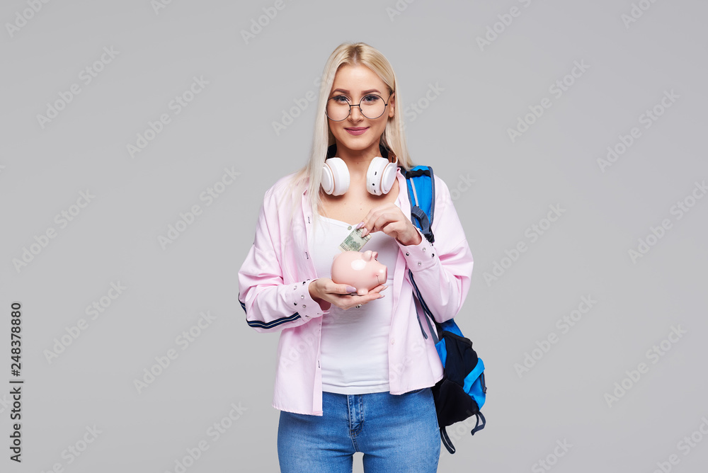 Cost of college education. Portrait of a happy female student with piggy bank and cash money in hands. The girl saves money for training in high school .Neutral grey background. Space for text Cost of college education. Portrait of a happy female student with piggy bank and cash money in hands. The girl saves money for training in high school .Neutral grey background. Space for text