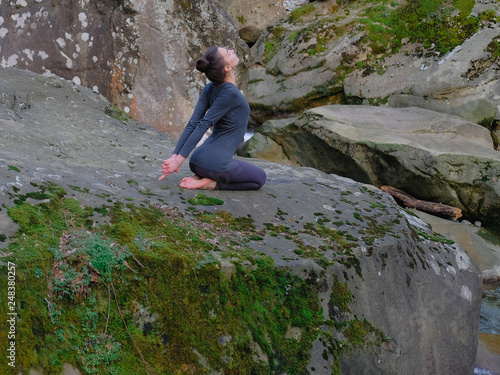 Young slim woman practicing yoga outdoors on big moss rock. Unity with nature concept. Girl sitting back bend