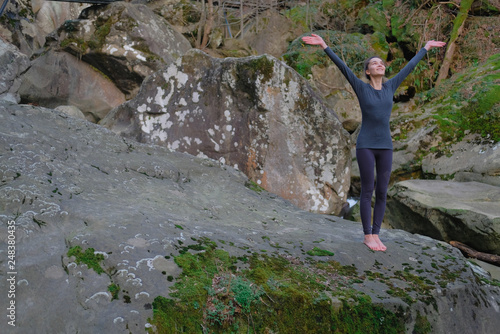 Young slim woman practicing yoga outdoors on big moss rock. Unity with nature concept. Girl standing with arms up