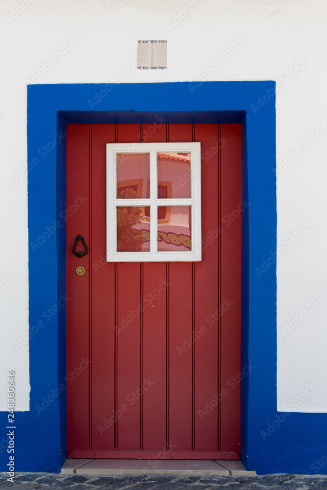 Red wooden door with four small windows. White wall with blue painting around the door. Number plate above the door. Red wooden door with four small windows. White wall with blue painting around the door. Number plate above the door.