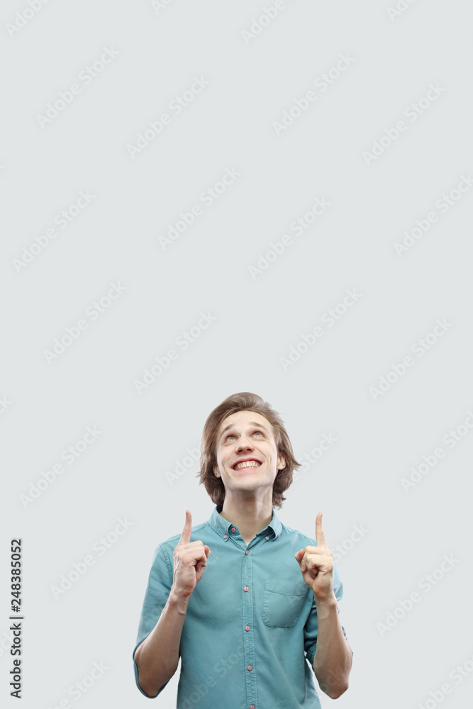 Portrait of happy toothy smiley handsome long haired blonde young man in blue casual shirt standing and pointing at up copyspace. indoor studio shot, isolated on light grey background. Portrait of happy toothy smiley handsome long haired blonde young man in blue casual shirt standing and pointing at up copyspace. indoor studio shot, isolated on light grey background.