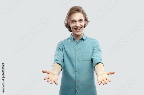 Φωτογραφία Portrait of happy genial handsome long haired blonde young man in blue casual shirt standing with raised arms and toothy smile looking at camera