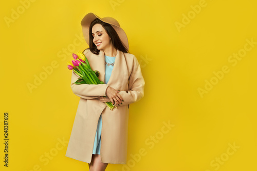 Fashion photo of a beautiful young woman with tulips in her hand .she dressed in a beautiful coat,  and T-shirt with stripes on the yellow background .Spring concept. March 8. beautiful girl.