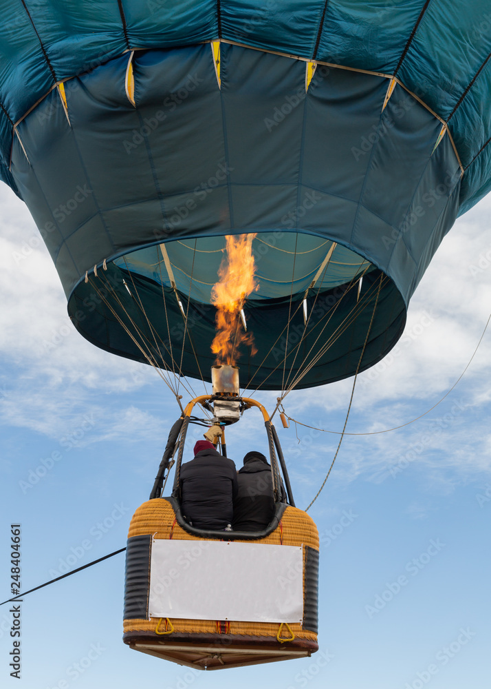 Two people ride in the basket of a hot air balloon as the flame from the  burner heats the air inside. A sign on the basket is blank. Stock Photo |  Adobe
