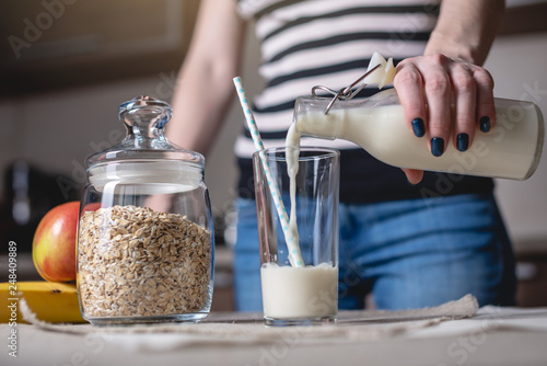Woman pours organic oat milk from a bottle into a glass on a table in the kitchen. Diet healthy vegetarian product