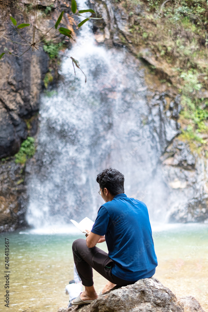Obraz premium Young man sit and turn back reading book at waterfall.