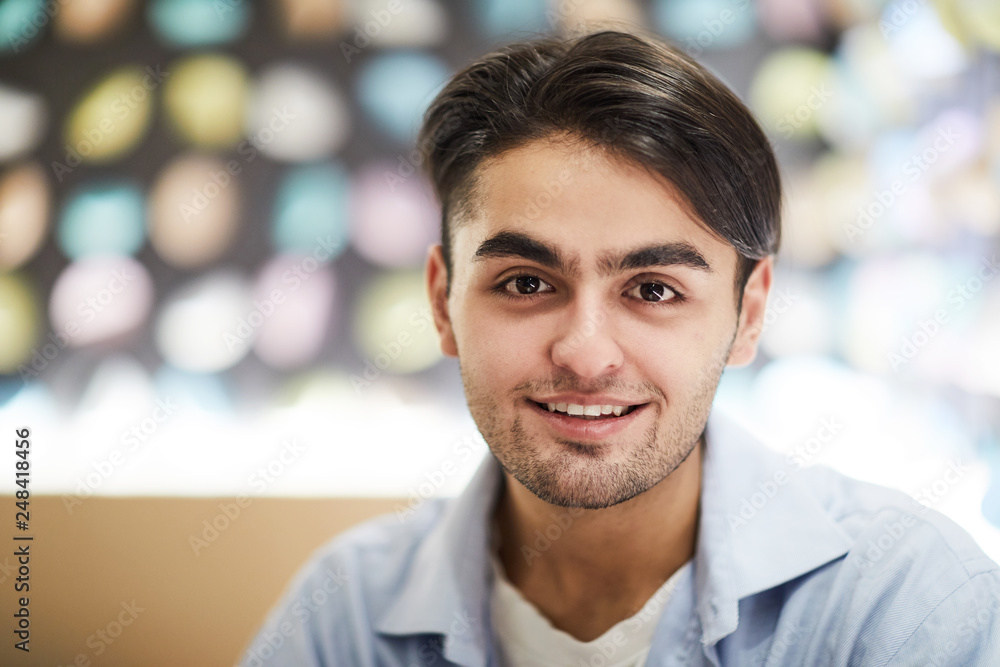 Young smiling handsome man in light blue shirt looking at you while sitting in front of camera in cafe Young smiling handsome man in light blue shirt looking at you while sitting in front of camera in cafe