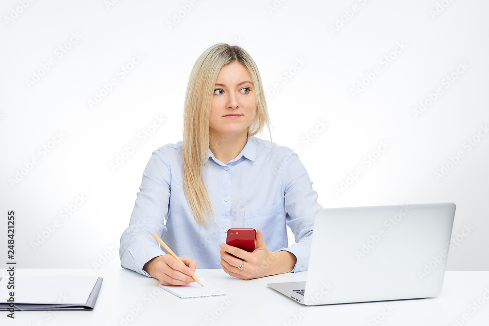 Young blonde woman looks away holds her red glass cell phone, holds a pencil and sits by the desk in the office with the white background. Aluminum notebook computer and documents on the table. Young blonde woman looks away holds her red glass cell phone, holds a pencil and sits by the desk in the office with the white background. Aluminum notebook computer and documents on the table.