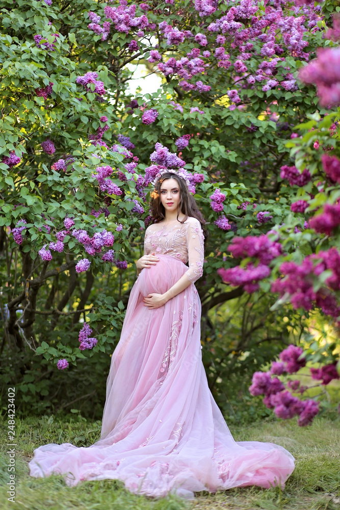 Beautiful young pregnant woman in a luxurious pink dress walking in a park of blossoming lilacs.