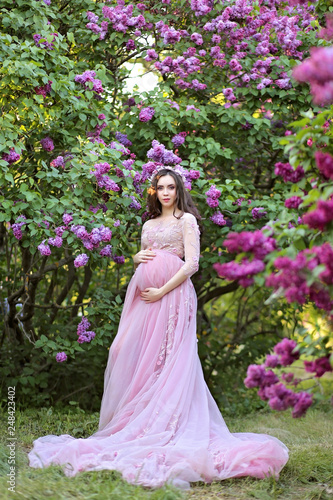 Beautiful young pregnant woman in a luxurious pink dress walking in a park of blossoming lilacs.