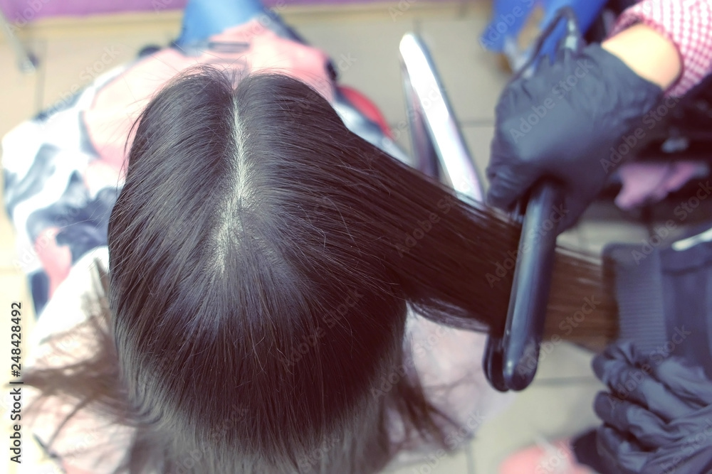 Fototapeta premium Hairdresser straights dark brown hair of beautiful woman using hair tongs in beauty salon. Back view.