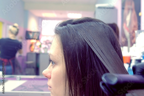 Hairdresser straights dark brown hair of beautiful woman using hair tongs in beauty salon. Side view.