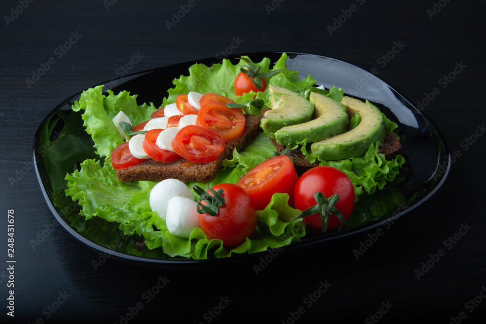 Salad and mozzarella, healthy sandwiches from black unleavened rye bread with fresh tomatoes and mozzarella cheese, with ripe avocados on a glossy black plate. Salad and mozzarella, healthy sandwiches from black unleavened rye bread with fresh tomatoes and mozzarella cheese, with ripe avocados on a glossy black plate.