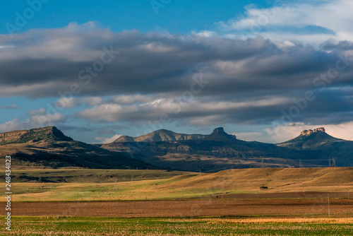 The Amphitheatre and surrounding mountains under a blue and cloudy sky with a valley of colorful plains lit up by the late evening sun. Drakensberg, South Africa