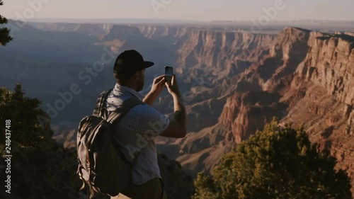 Slow motion happy young tourist man with backpack taking smartphone photo of epic summer sunset over Grand Canyon USA.