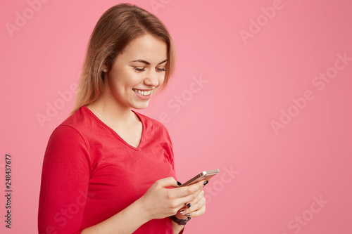 Smiling pleasant looking woman in red outfit holds smart phone, searches social networks, has short hairstyle, connected to wireless internet, isolated over rosy background with empty space for text