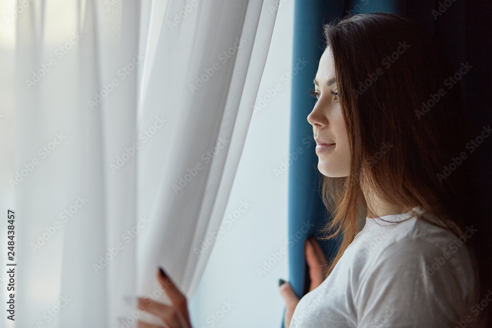 Horizontal shot of attractive brunette pensive young woman in casual white t shirt, looks out of window with thoughtful expression, has make up, enjoys calm domestic atmosphere, poses at her flat. Horizontal shot of attractive brunette pensive young woman in casual white t shirt, looks out of window with thoughtful expression, has make up, enjoys calm domestic atmosphere, poses at her flat.