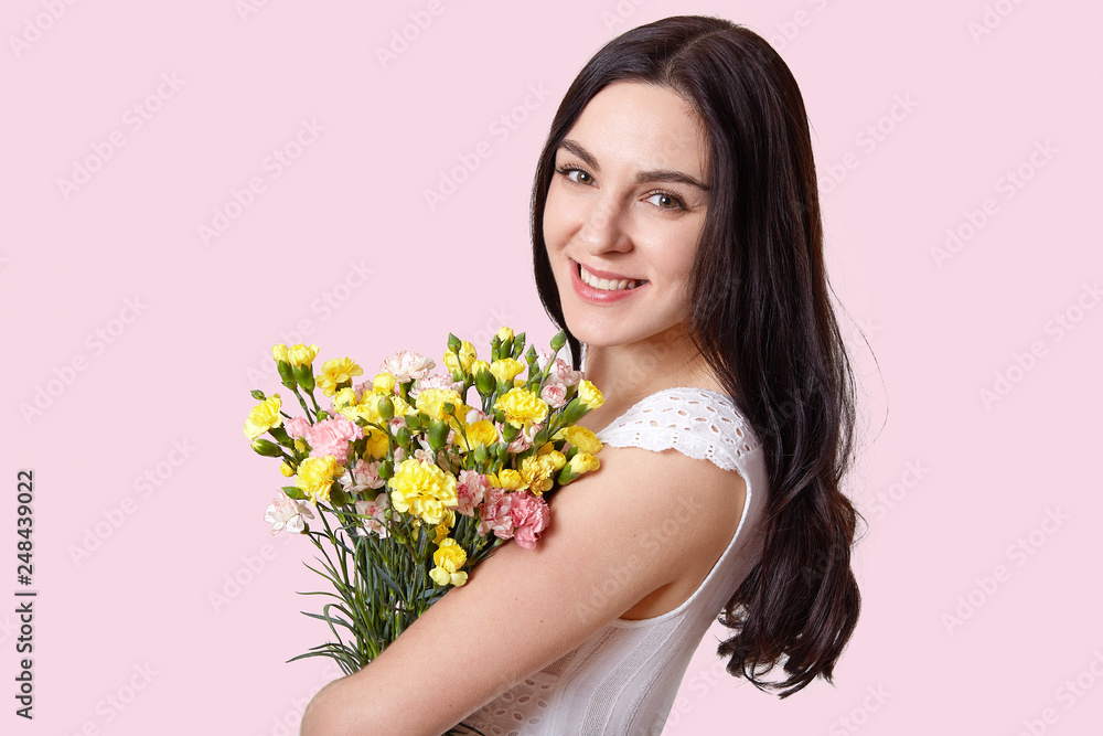 Close up shot of attractive woman with make up, natural beauty, gentle smile, has long straight hair, carries flowers, recieves bouquet on 8 March, wears stylish dress, isolated over rosy background Close up shot of attractive woman with make up, natural beauty, gentle smile, has long straight hair, carries flowers, recieves bouquet on 8 March, wears stylish dress, isolated over rosy background
