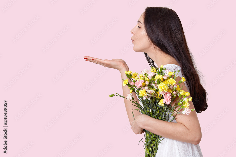 Profile shot of brunette European woman blows air kiss, stretches palm, expresses love, holds flowers, dressed in white dress, poses over light pink background with copy space for your promotion Profile shot of brunette European woman blows air kiss, stretches palm, expresses love, holds flowers, dressed in white dress, poses over light pink background with copy space for your promotion