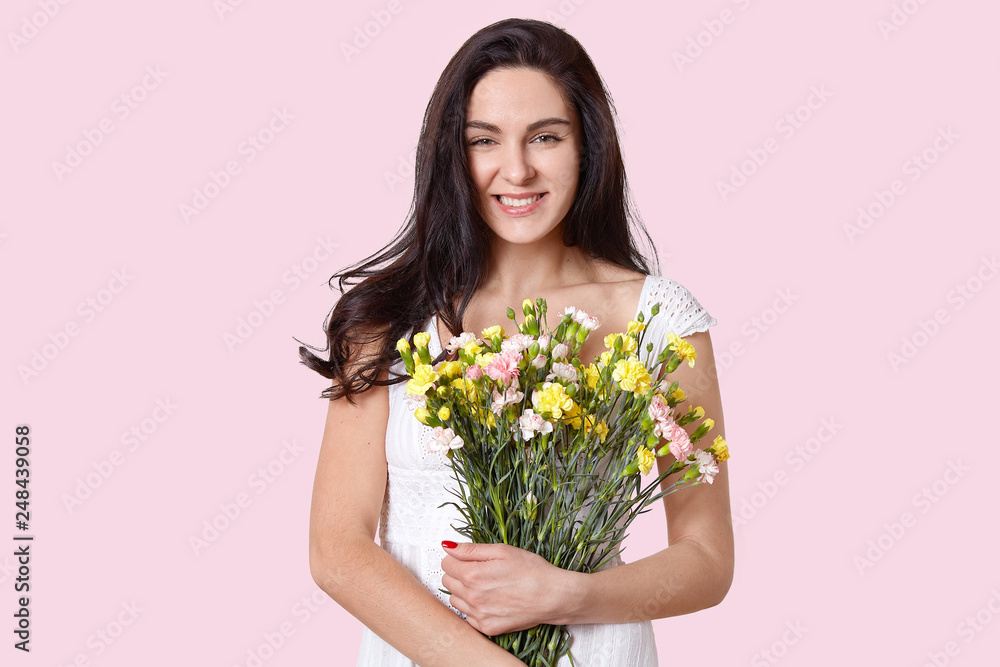 Sensual woman with toothy smile, has long straight hair, holds flowers closely, dressed in white dress, enjoys spring time and holiday, poses over rosy background, expresses positive emotions Sensual woman with toothy smile, has long straight hair, holds flowers closely, dressed in white dress, enjoys spring time and holiday, poses over rosy background, expresses positive emotions