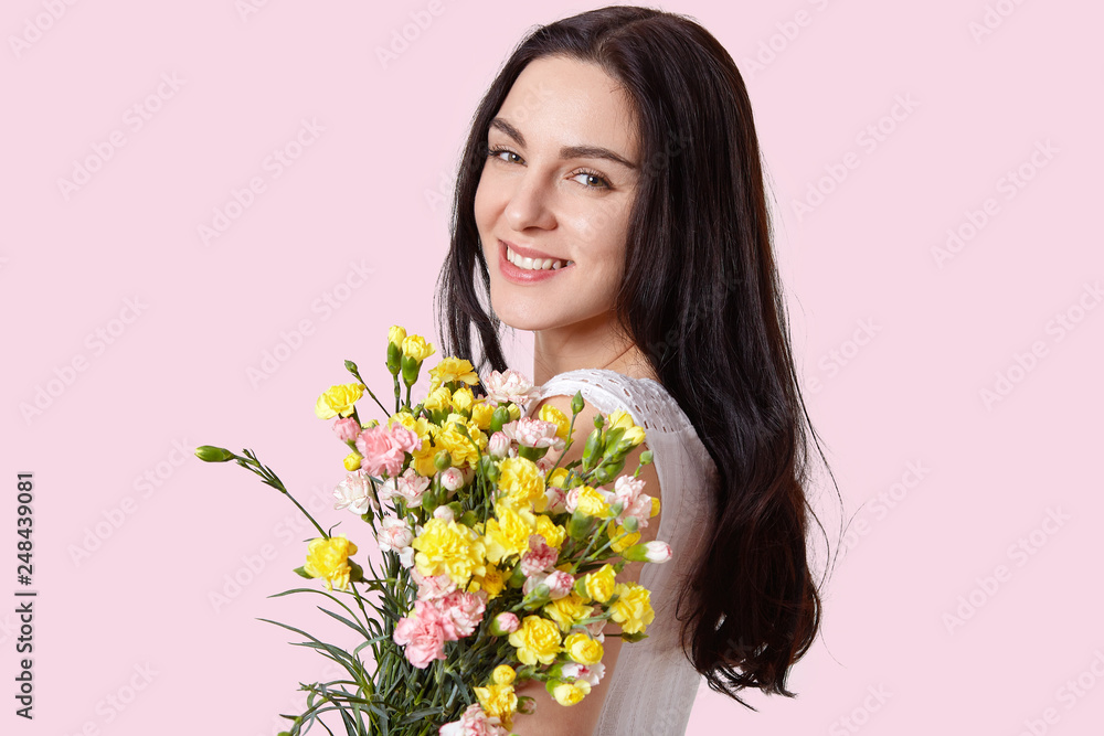 Headshot of pretty European young woman with tender smile, healthy skin, dark long hair, carries bouquet of spring flowers, looks at camera with happiness, isolated over light pink background Headshot of pretty European young woman with tender smile, healthy skin, dark long hair, carries bouquet of spring flowers, looks at camera with happiness, isolated over light pink background
