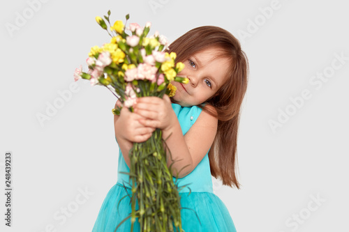 Horizontal shot of pretty dark haired female child in festive dress stretches hands with beautiful flowers  has appealing look  isolated over white background. Children  flowers  holiday concept