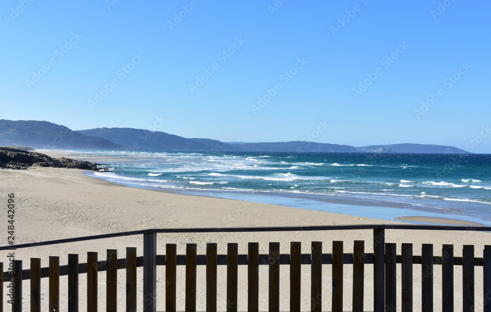 Wild beach with wooden handrail, golden sand and blue sea with waves. Sunny day, clear sky. Galicia, Coruña Province, Spain. Wild beach with wooden handrail, golden sand and blue sea with waves. Sunny day, clear sky. Galicia, Coruña Province, Spain.